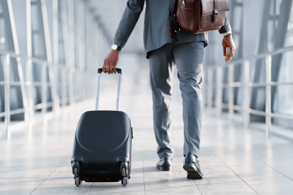 Man walking down a glass hallway in an airport pulling along a rolling carry-on suitcase